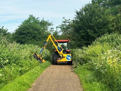 Yellow and blue tractor with extended boom arm working on a vegetation-lined pathway on a sunny day